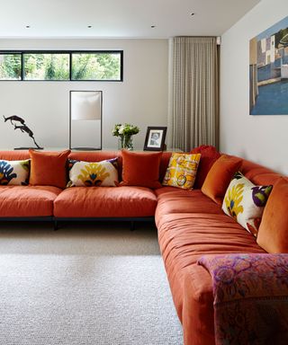 Large family room with orange wall to wall corner sofa and selection of printed cushions, cream wool carpet, small panel windows