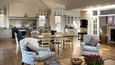 A cozy kitchen space with a large dining table and seating area with patterned blue and white upholstered chairs. The cabinets are white, with a tiled backsplash and large black AGA oven, and the flooring is wood.