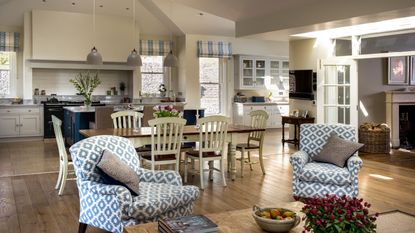 A cozy kitchen space with a large dining table and seating area with patterned blue and white upholstered chairs. The cabinets are white, with a tiled backsplash and large black AGA oven, and the flooring is wood.