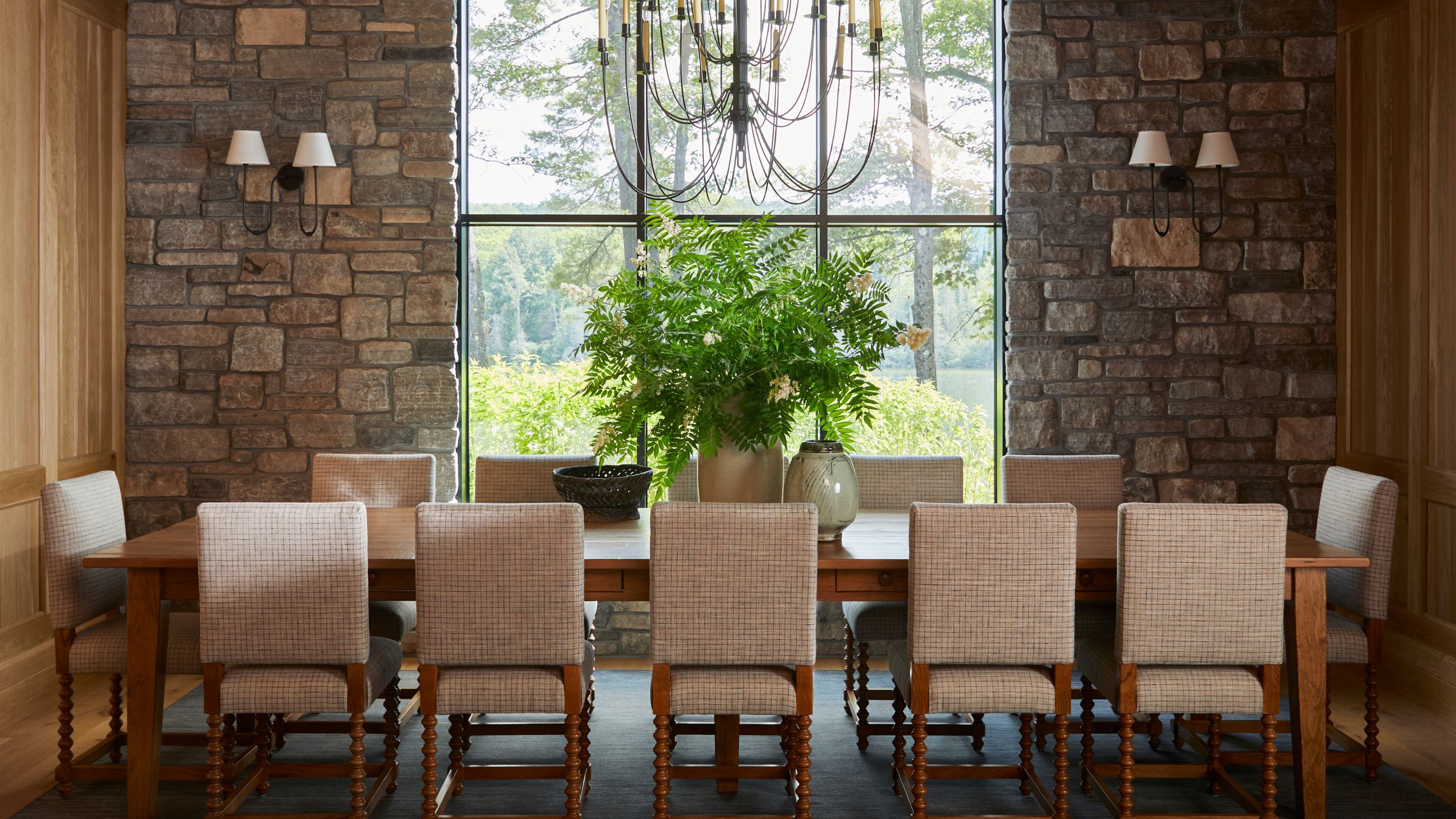 A long wooden dining table with high-back upholstered chairs set against a textured stone wall and a large window overlooking the woods.