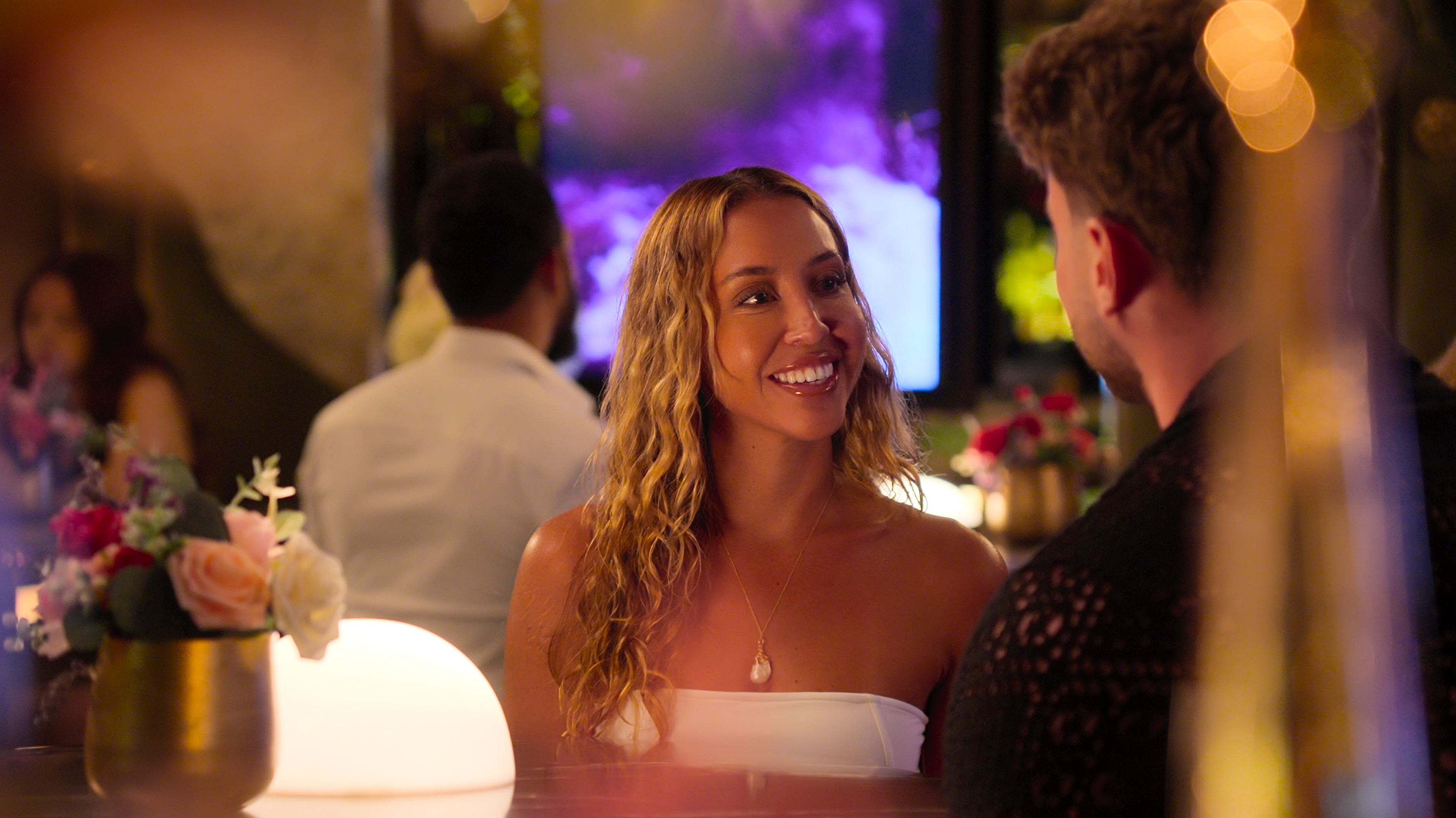 Amber Mozo sets with Harry Jowsey (unseen) at a restaurant bar with golden drinkware and bottles blurred in the frame, in Netflix's 'Let's Marry Harry.'