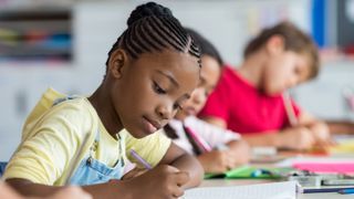 A Black girl writes in a notebook in a classroom.