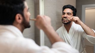 A man with dark hair and wearing a white dressing gown brushes his teeth before bed