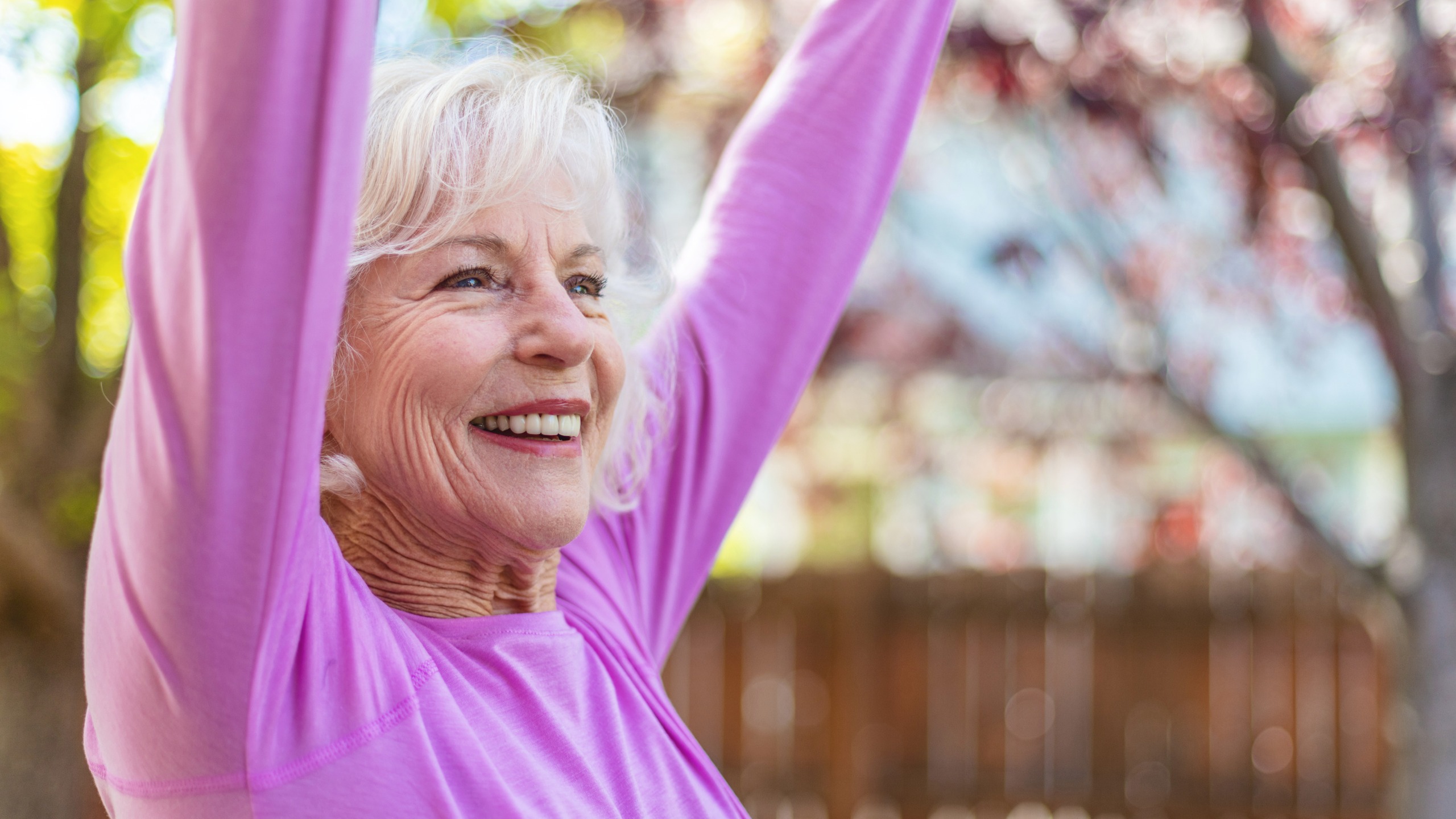 A woman in a bright pink top stands outside smiling with her arms stretched to the sky