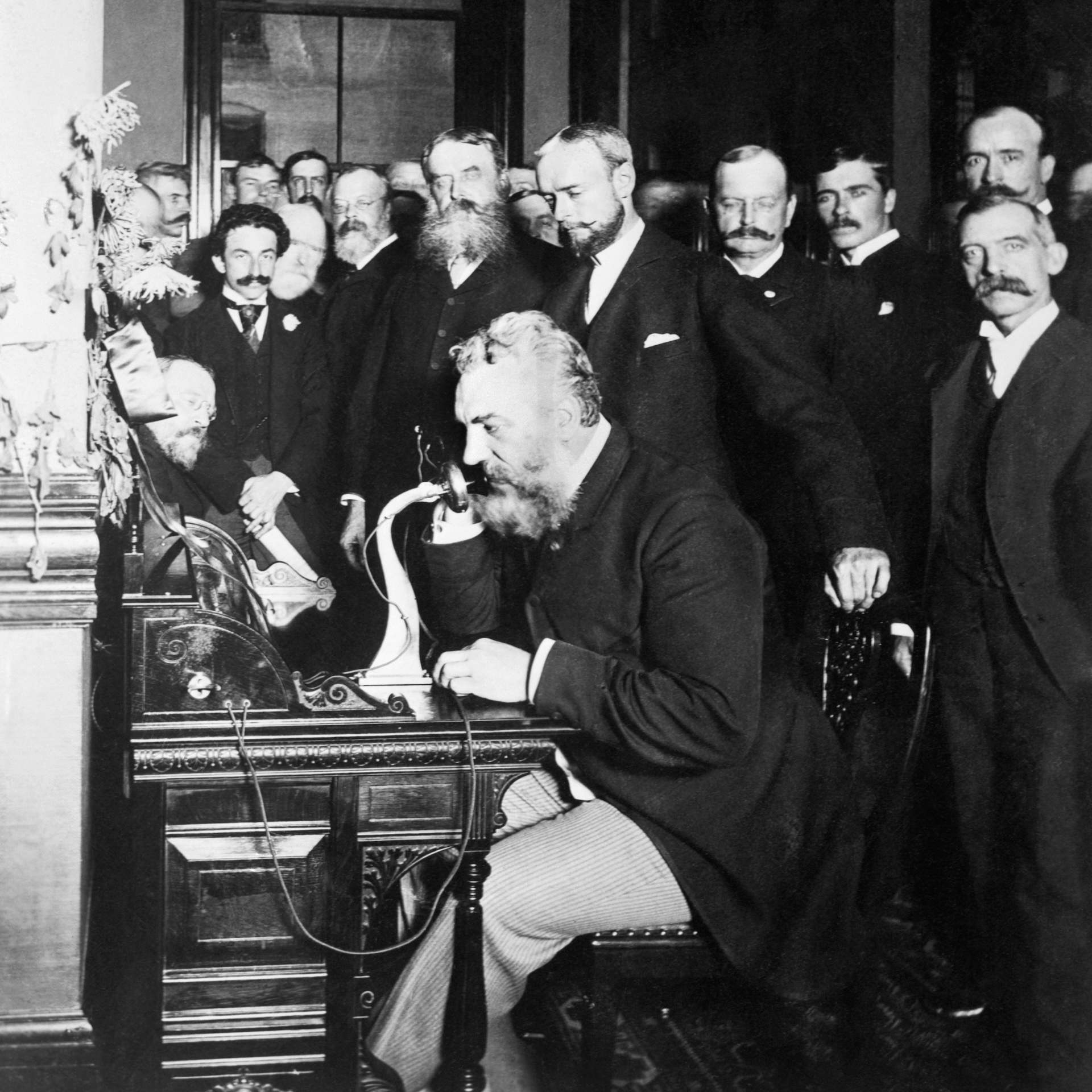 a black-and-white photograph of a group of businessmen standing behind Alexander Graham Bell as he speaks on a telephone