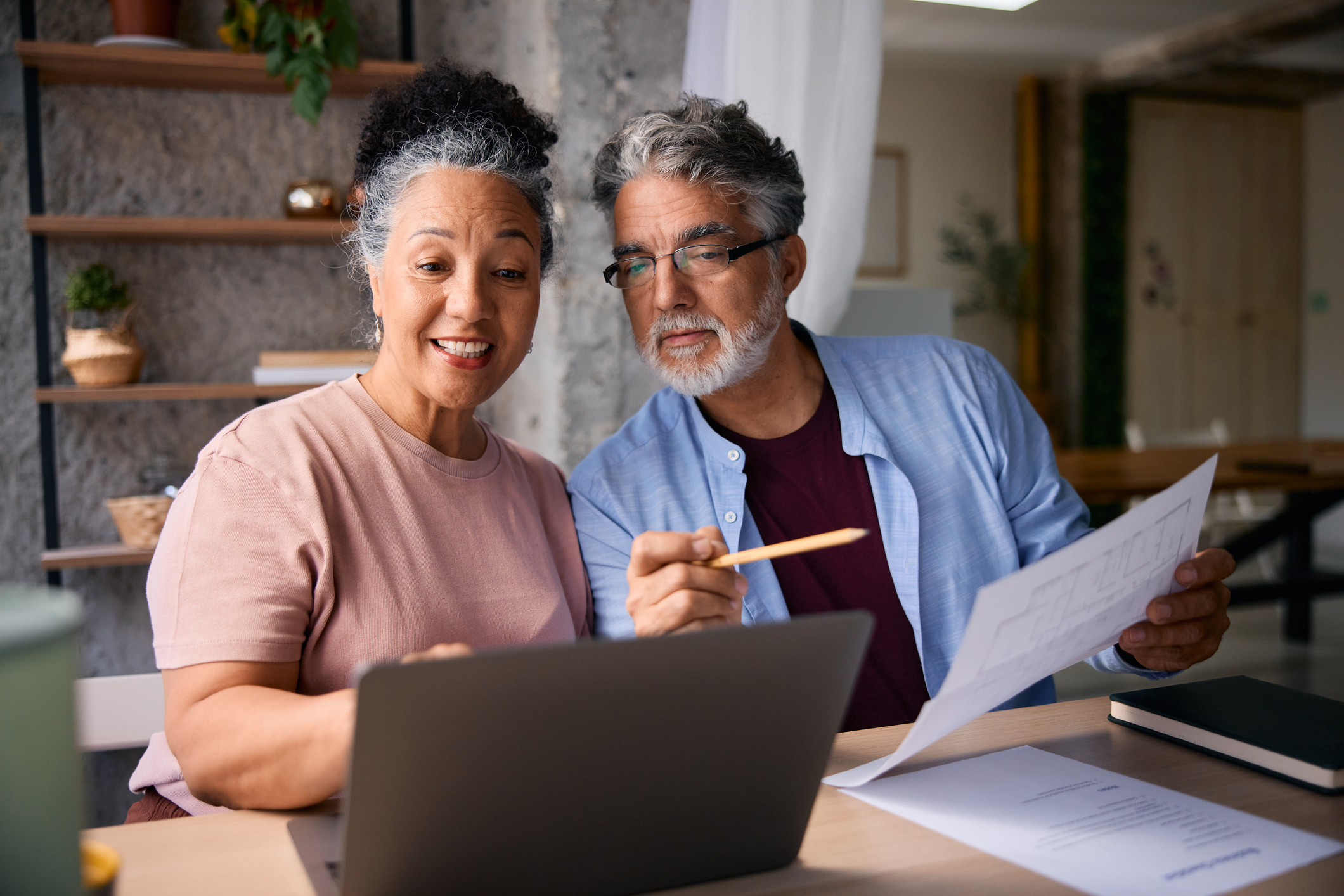 Senior couple sitting together at a table, calculating domestic bills while using a laptop and reviewing documents, managing their finances and enjoying their time at home