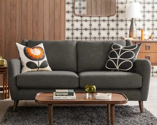 A grey mid-century sofa with Orla Kiely floral scatter cushions, grey wallpaper and brown wood wall paneling