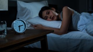 A woman with dark brown hair lies on her side staring at the back of a white alarm clock that shows the time as 2.30am