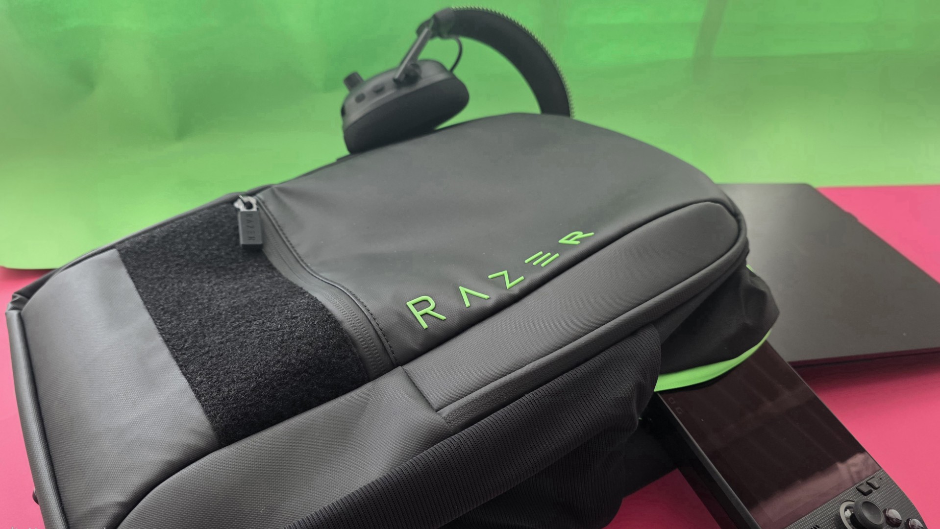 A black Razer backpack, Razer headphones, a black laptop, are placed on a split pink and green background.