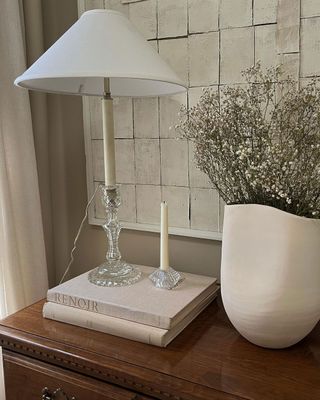 Glass decorative lamp with white shade sits upon small elegantly displayed books next to white ceramic vase with delicate white small petaled flowers