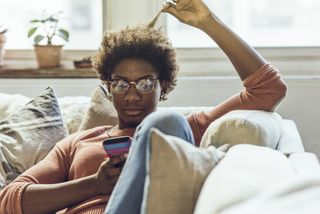 woman sitting on sofa at home, looking at her phone and twiddling her hair with the other hand