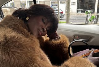 Amy in a car wearing a fur coat, black bag, and gold earrings.