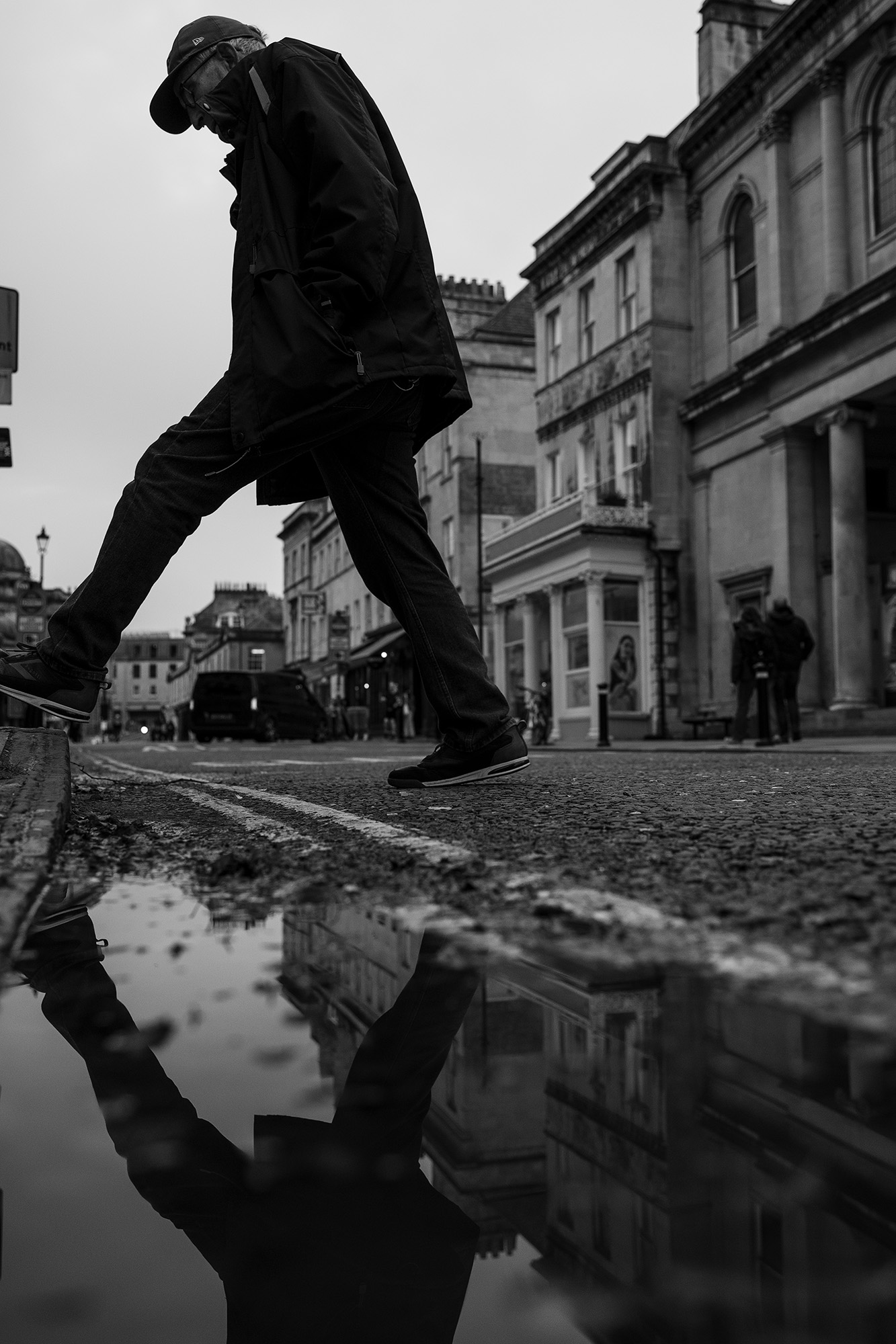 Black and white street photograph of man stepping over puddle