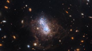 The dwarf irregular galaxy I Zwicky 18 lies at the center of this image. The bright region of white and blue stars at the galaxy’s core appear as two distinct lobes, representing different periods of star formation. 
