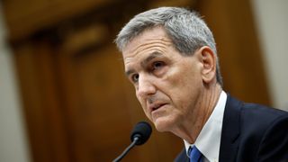 a man in a suit speaks into a microphone in a large hearing room