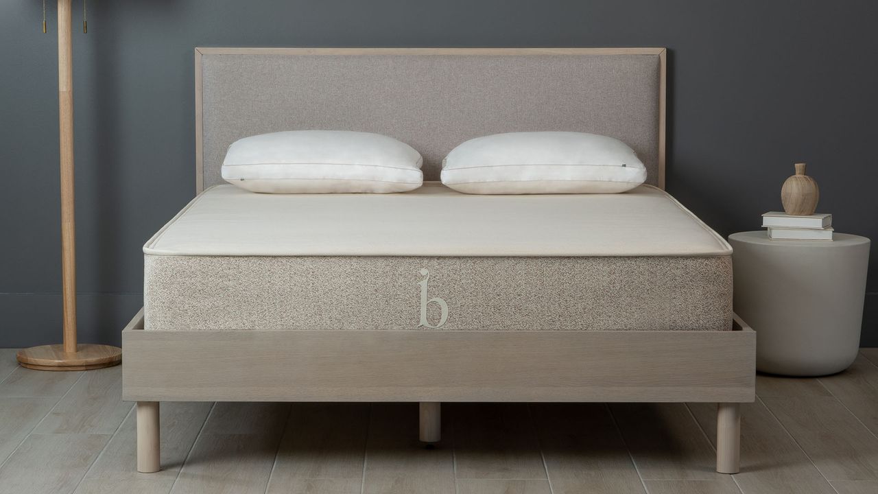 Birch Natural mattress on a neutral wooden bedframe against a dark wall, with a lamp and bedside table