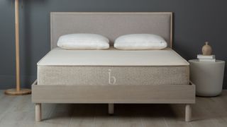 Best mattress in a box: Birch Natural mattress on a neutral wooden bedframe against a dark wall, with a lamp and bedside table