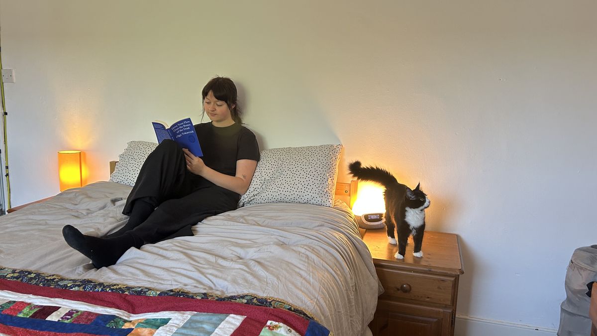 The DreamCloud Luxe Memory Foam mattress dressed in grey bedding with a colorful throw. Our tester sits and reads a book while her cat stands on the bedside table next to her