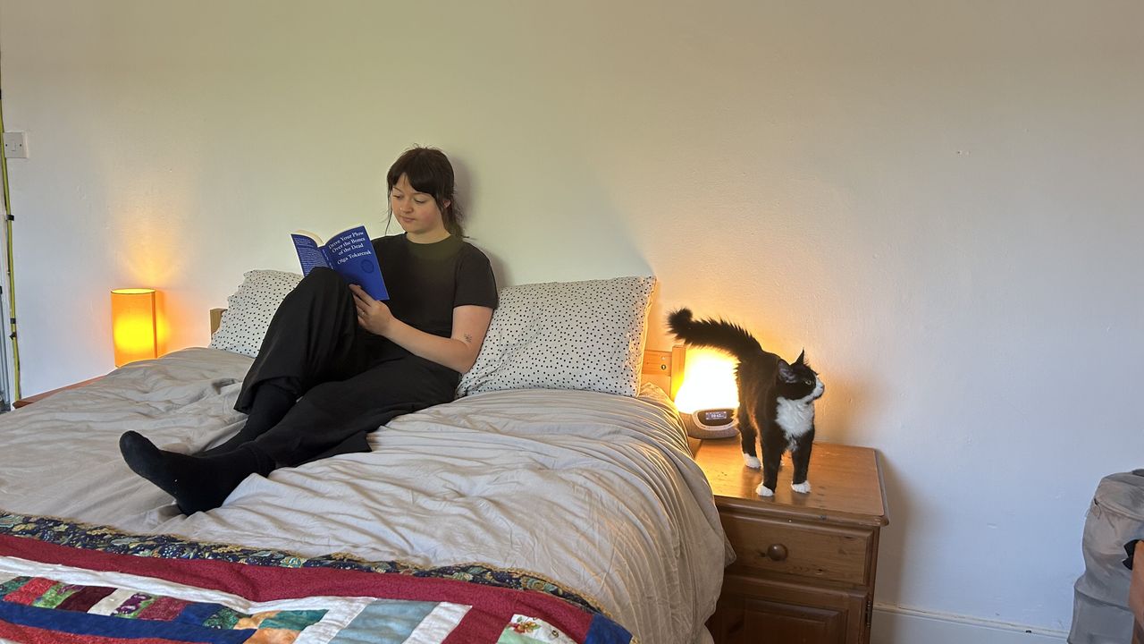 The DreamCloud Luxe Memory Foam mattress dressed in grey bedding with a colorful throw. Our tester sits and reads a book while her cat stands on the bedside table next to her