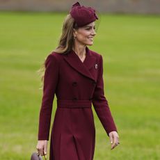 Kate Middleton walks outside on grass wearing a burgundy coat with chocolate brown heels. She carries a burgundy Chanel bag.