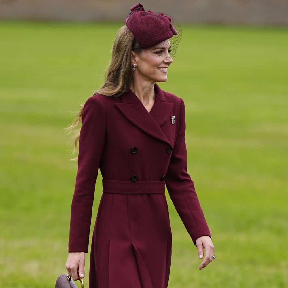 Kate Middleton walks outside on grass wearing a burgundy coat with chocolate brown heels. She carries a burgundy Chanel bag.