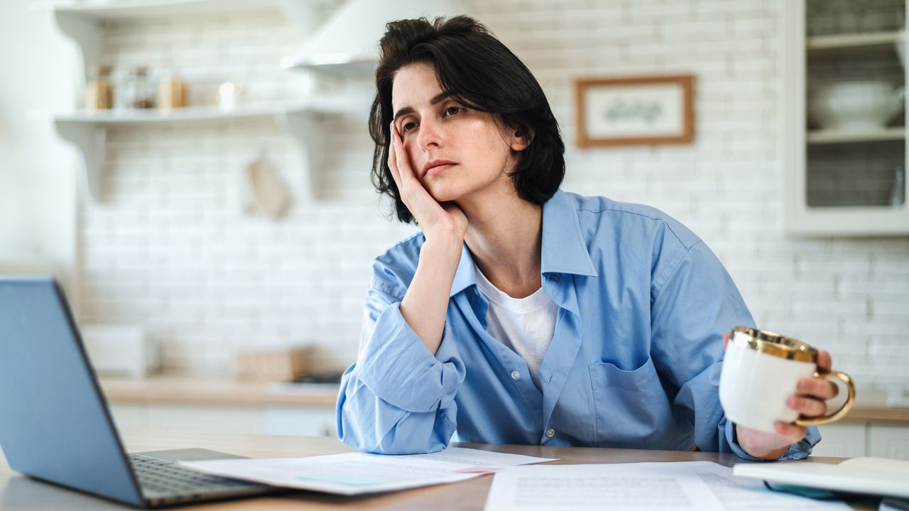 A tired-looking woman holds a coffee cup and looks at her laptop in her kitchen.