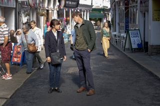 A still from episode one of Beyond Paradise showing DS Esther Williams (Zahra Ahmadi) and DI Humphrey Goodman (Kris Marshall) standing in the middle of Shipton Abbott's main shopping street. He has his hands in his pockets, and she is staring at him from beneath her fringe.