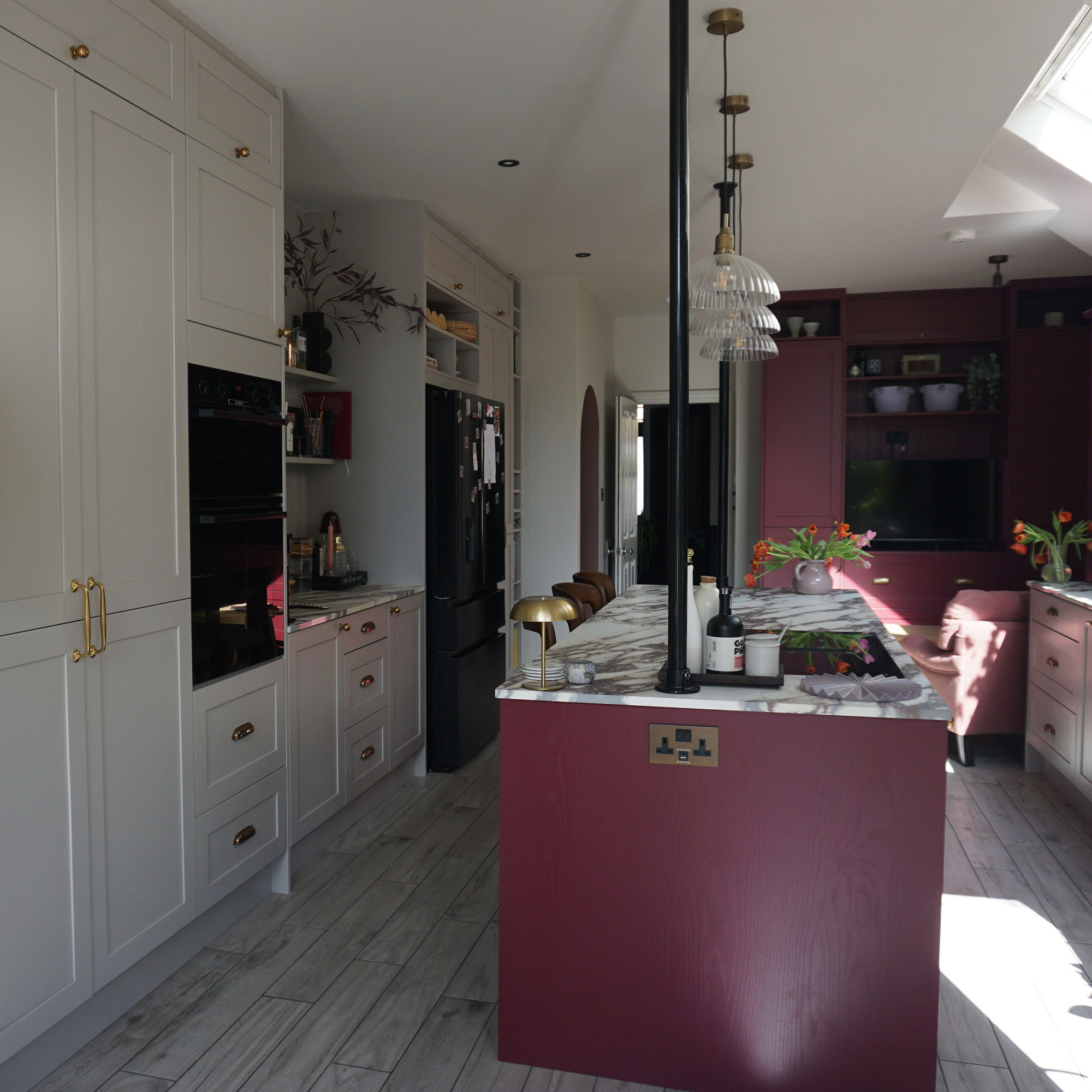 Kitchen island painted in burgundy, topped with marble, integrated brass plug, across from run of light grey cabinetry