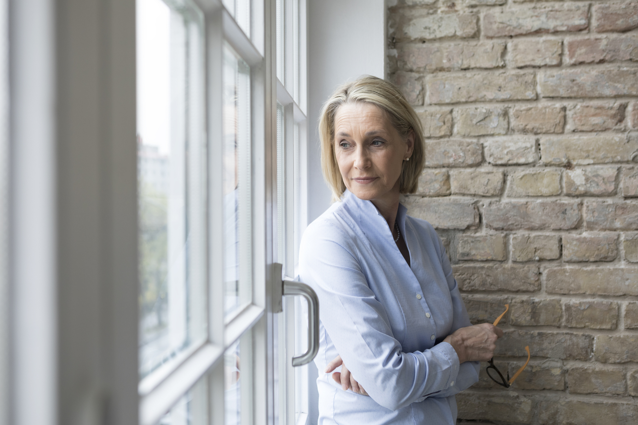 A woman standing against a brick wall, looking out the window.