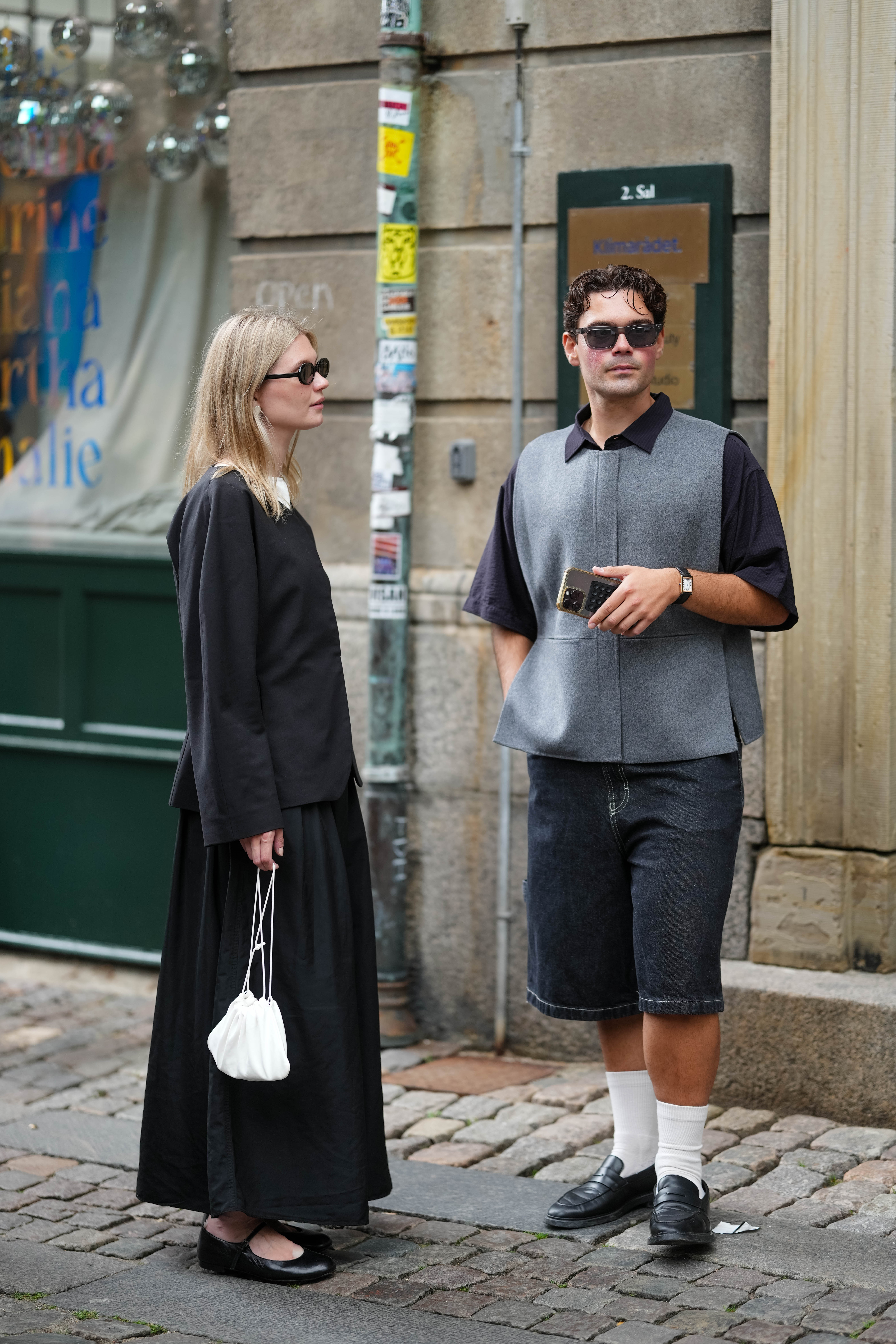 COPENHAGEN, DENMARK - AUGUST 04: A guest (L) wears a black long-sleeve top with a round neckline, paired with a flowing black maxi skirt. Accessories include black rectangular sunglasses and a small white drawstring pouch bag. Footwear consists of black flat shoes. Hair is styled straight and loose, a guest (R) wears black sunglasses, a black short sleeves oversized t-shirt with a gray sleeveless jacket, dark gray denim bermuda knees shorts, white socks, black shiny leather loafer, a black shiny leather watch, outside Forza Collective, during Copenhagen Fashion Week, on August 04, 2025 in Copenhagen, Denmark (Photo by Edward Berthelot/Getty Images)