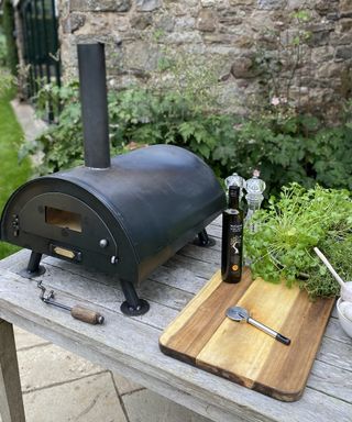 A table top pizza oven on grey wooden table with wooden board, pizza cutter and bottle of extra virgin olive oil in black glass bottle with screw top lid