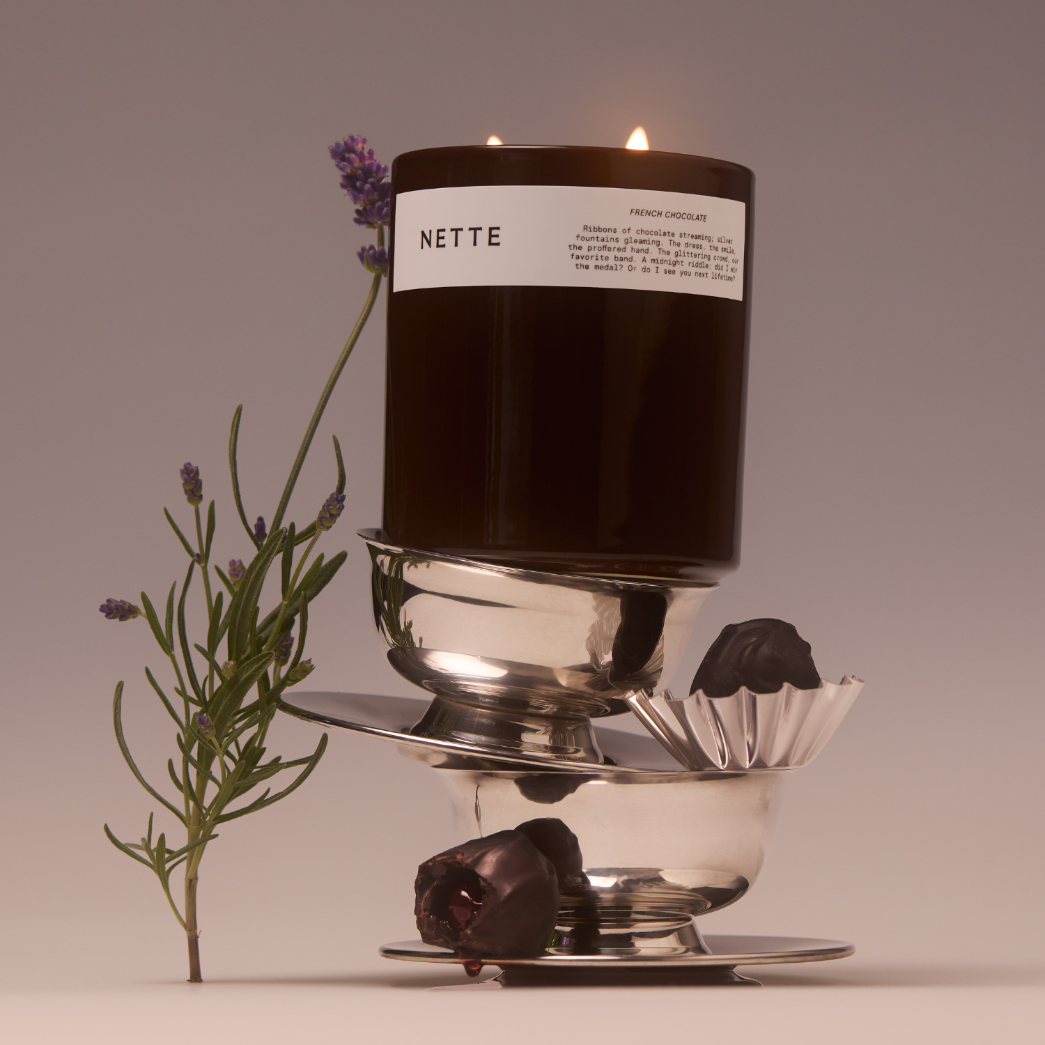 A brown glass candle on a stack of silver coupes by a sprig of lavender and a pair of chocolate truffles