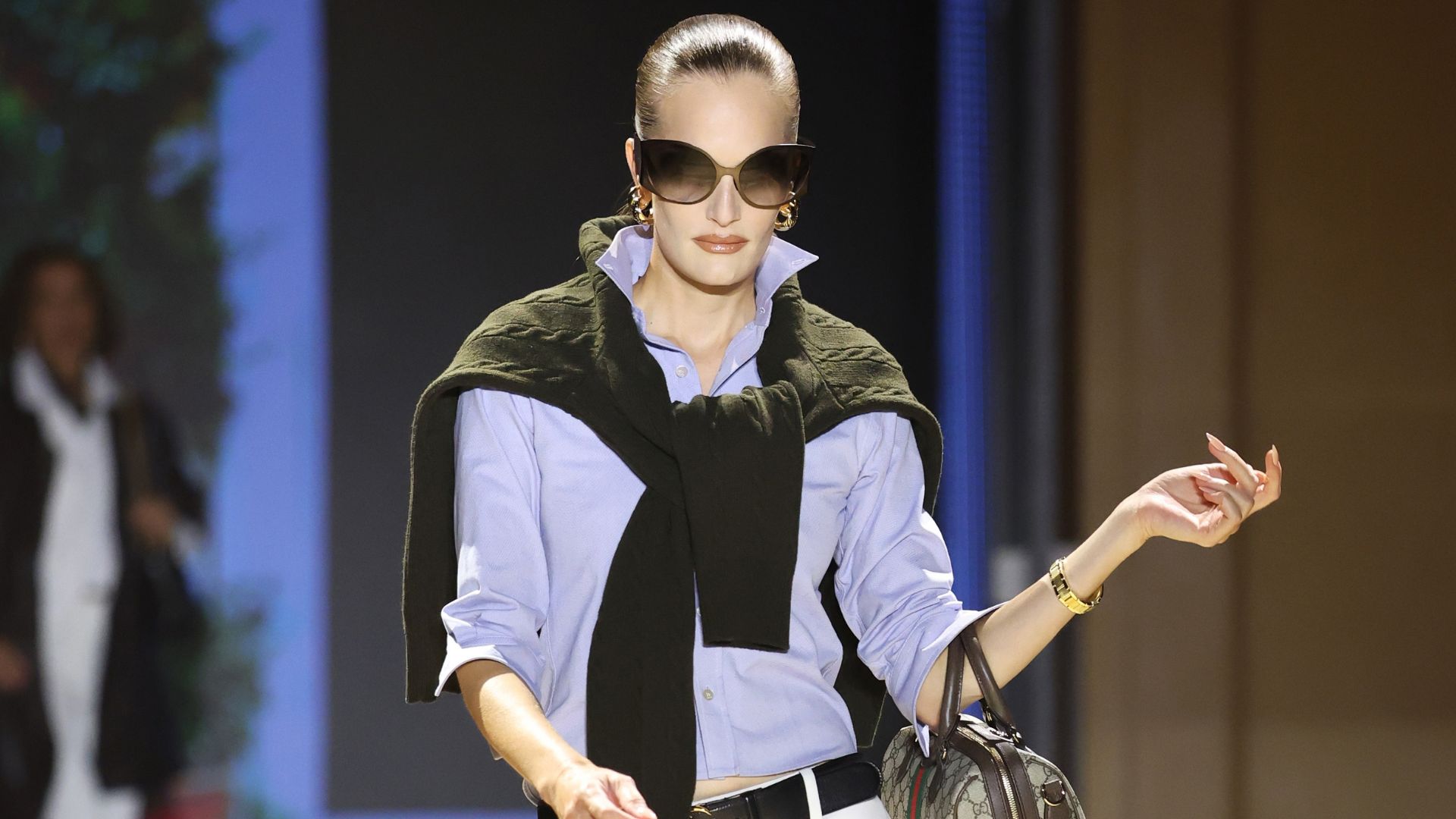 Model walks the Gucci SS27 Runway wearing jodhpur leggings tucked into a shirt with a jumper over the shoulder and black knee-high boots.