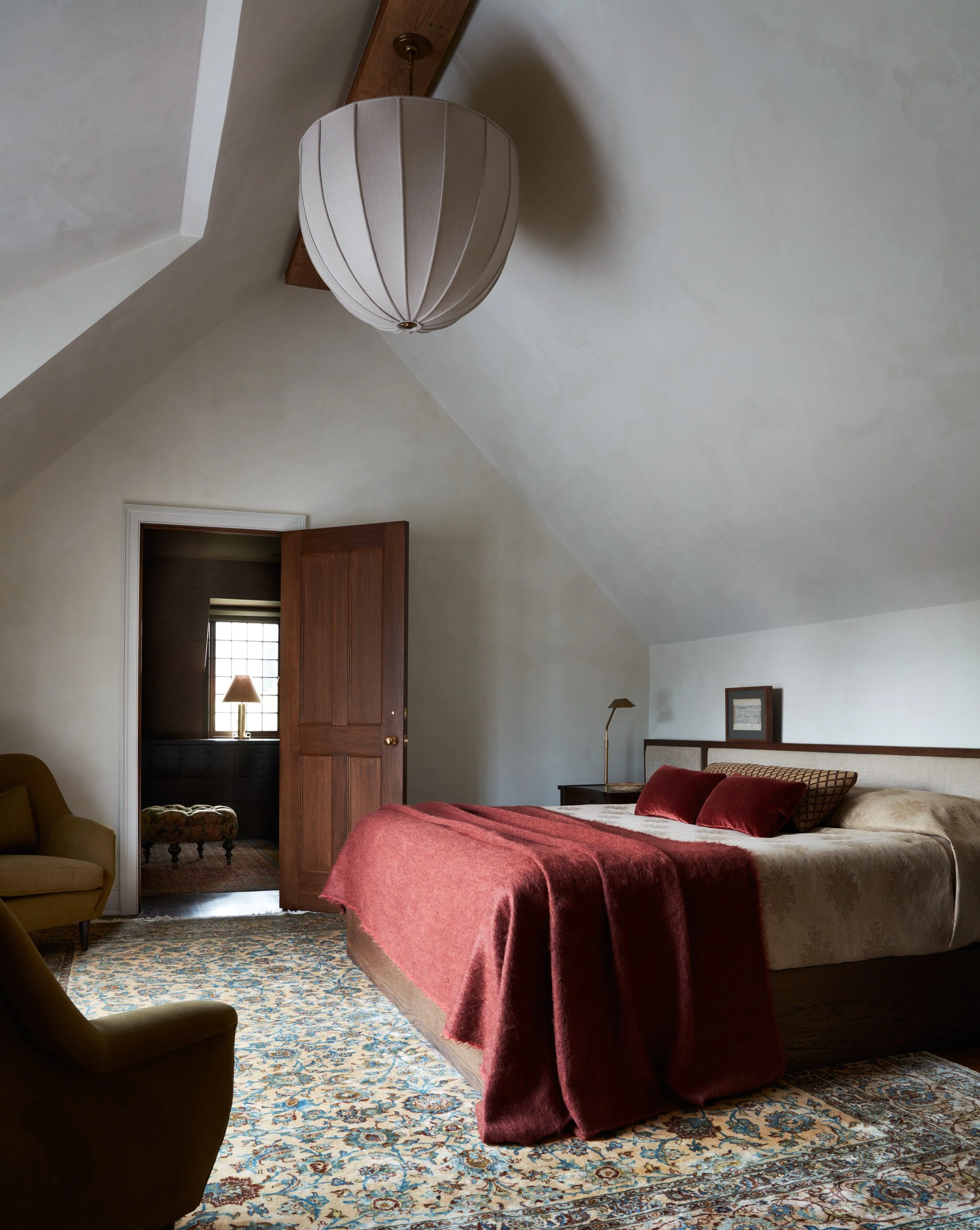 image of a traditional bedroom with angled white ceilings and white walls. There is a beige bed with an upholstered extended headboard and a wooden side table next to it. The bed has beige and red bedding. There is a blue and beige patterned rug on the ground.