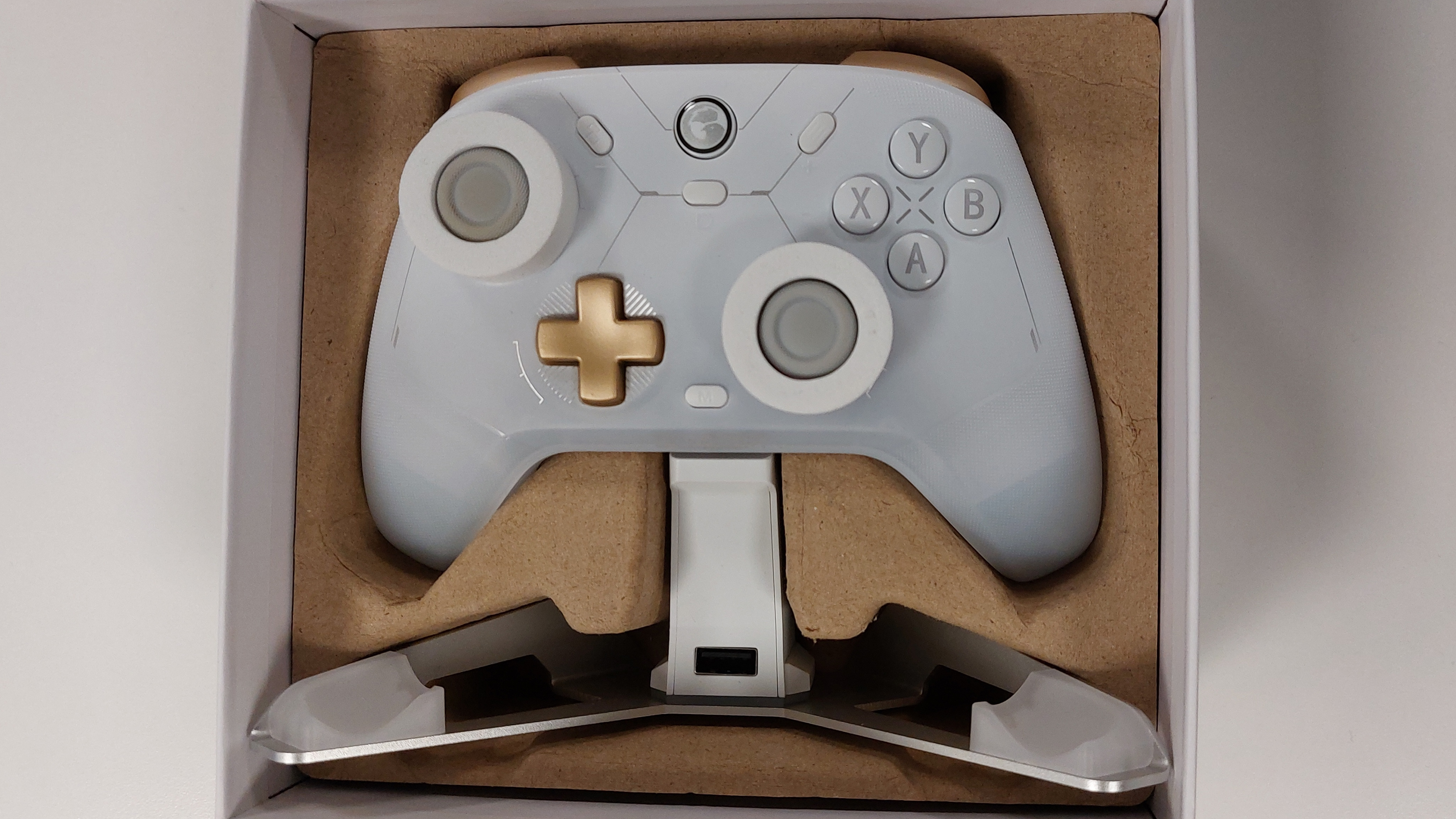 The GameSir Cyclone 2 controller in Phantom White seen wresting in the cardboard insert within its box. This colourway features a white gamepad body, a golden D-pad, golden shoulder buttons, grey thumbstick covers with orange thumbstick inlays (currently obscured by the styrofoam thumbstick guards the controller arrives with), and silver text accents. The RGB lights are switched off in this image. The charging cradle of the Charging Station edition is also present in the box, featuring a boomerang shaped stand with one central pillar to hold the controller aloft.