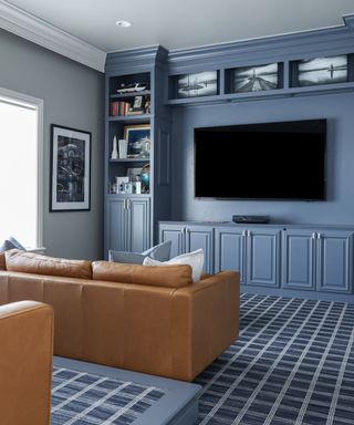 A small blue theater room with a bookcase around the black rectangular TV, a light brown leather couch facing it, and blue and white grid carpet on the floor