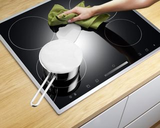 White woman's arm cleaning an electric stove top with a green cloth while pan comes to the boil