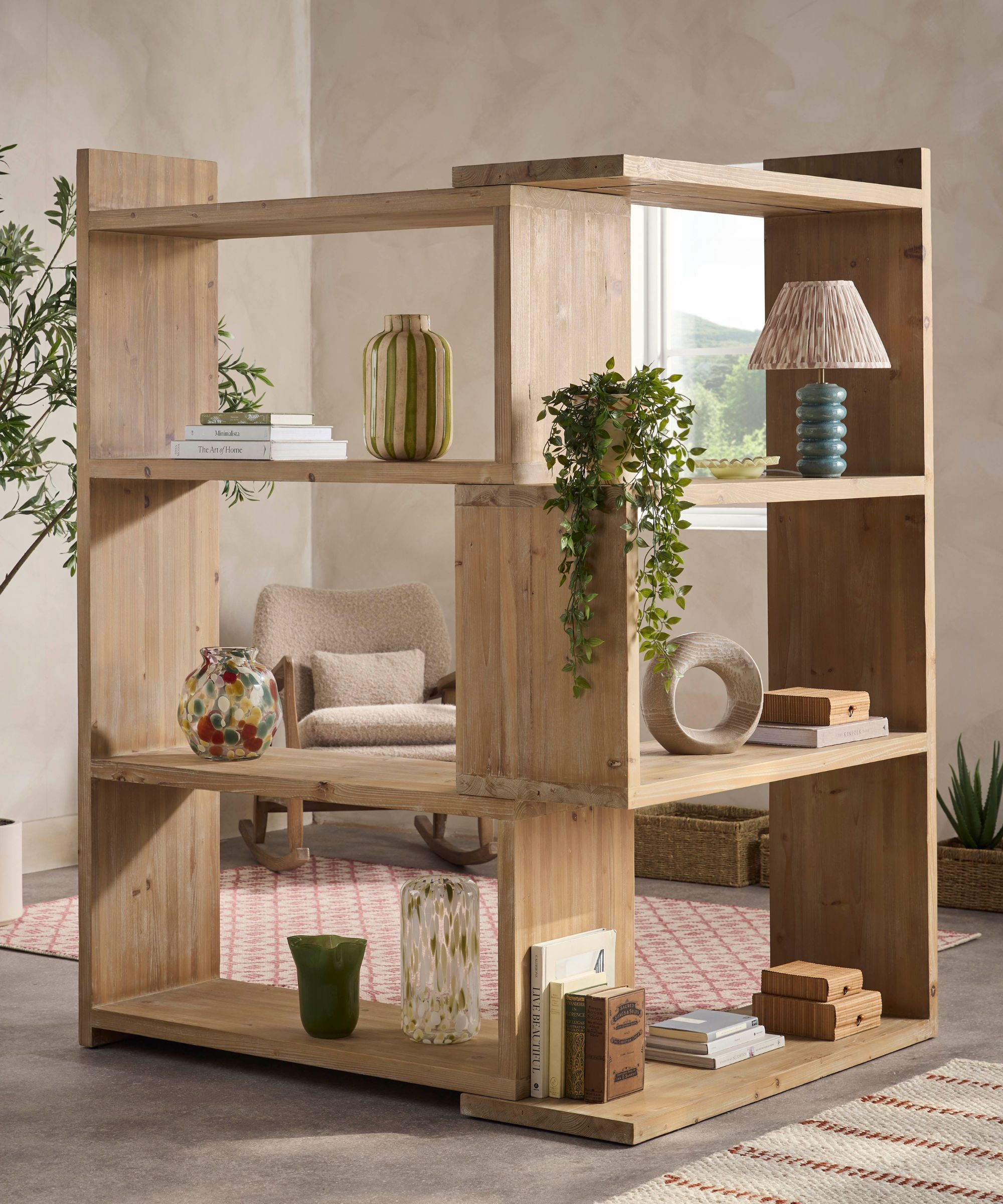 A freestanding open wooden shelving unit made of light natural timber, arranged in a grid of square compartments. The shelves display books, glass vases, a table lamp, trailing greenery, and small decorative objects. Through the open sections, a cozy armchair and patterned rug are visible behind.