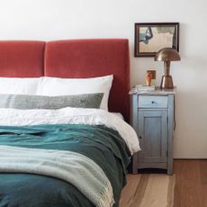 A bedroom with red velvet headboard and green and white bedding. A brushed brass lamp stands on a blue bedside cupboard and there's a piece of abstract art hanging on the wall