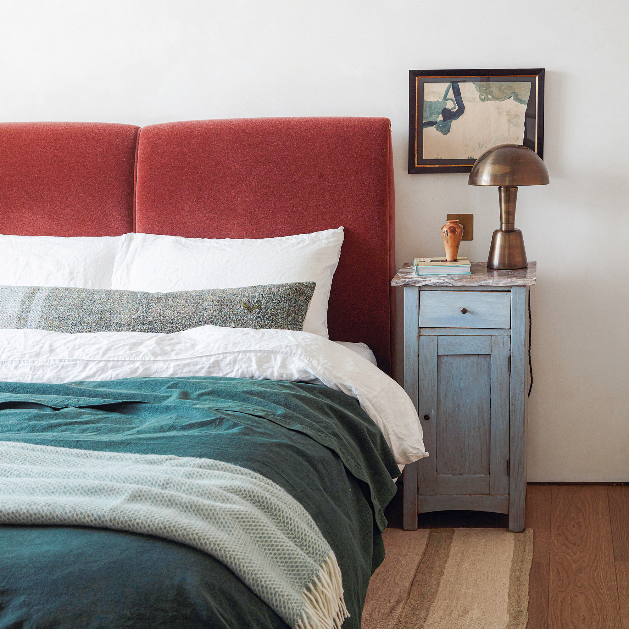 A bedroom with red velvet headboard and green and white bedding. A brushed brass lamp stands on a blue bedside cupboard and there's a piece of abstract art hanging on the wall