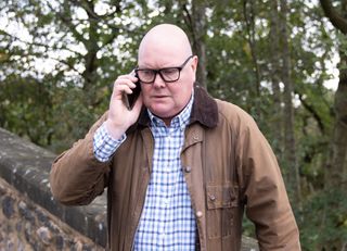 Paddy Kirk on the phone wearing a check shirt and brown jacket in Emmerdale