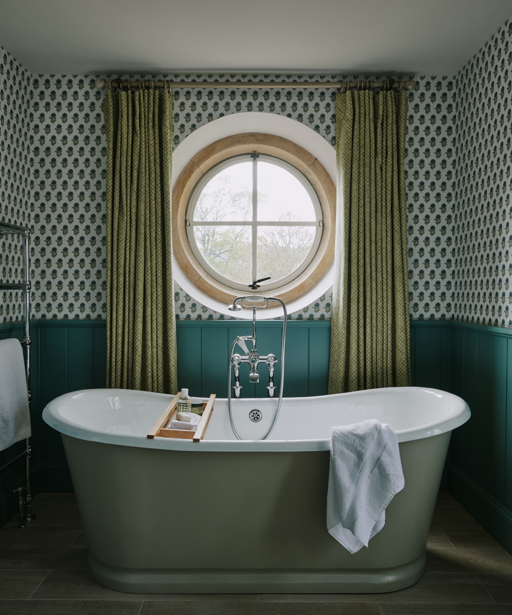 A bathroom with teal wall paneling on the lower walls, wallpaper on the upper walls, and a round mirror with green curtains. A green roll-top bath in the middle of the room.