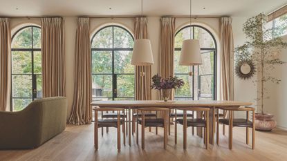 Open plan dining room with three large arched windows each with long cream drapes. A large wooden dining table sits in the middle of the space with two pendant lights with white shades hanging above