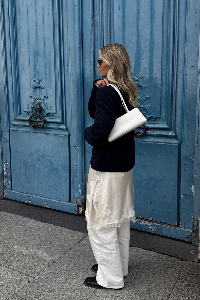 A sweater styling trend is shown in a photo of a woman&#039;s outfit, where she&#039;s standing outside in front of a blue doorway in Paris wearing a navy blue turtleneck sweater over a white slip dress and matching white wide leg jeans styled with black ankle boots and a white shoulder bag