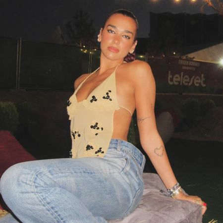 Dua Lipa sitting on hay wearing a printed halter top, jeans, and red Puma Speedcat sneakers.