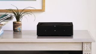A black Sonos Amp on a table surface, next to a small pot plant. On the wall behind, the corner of a wooden picture frame is visible.