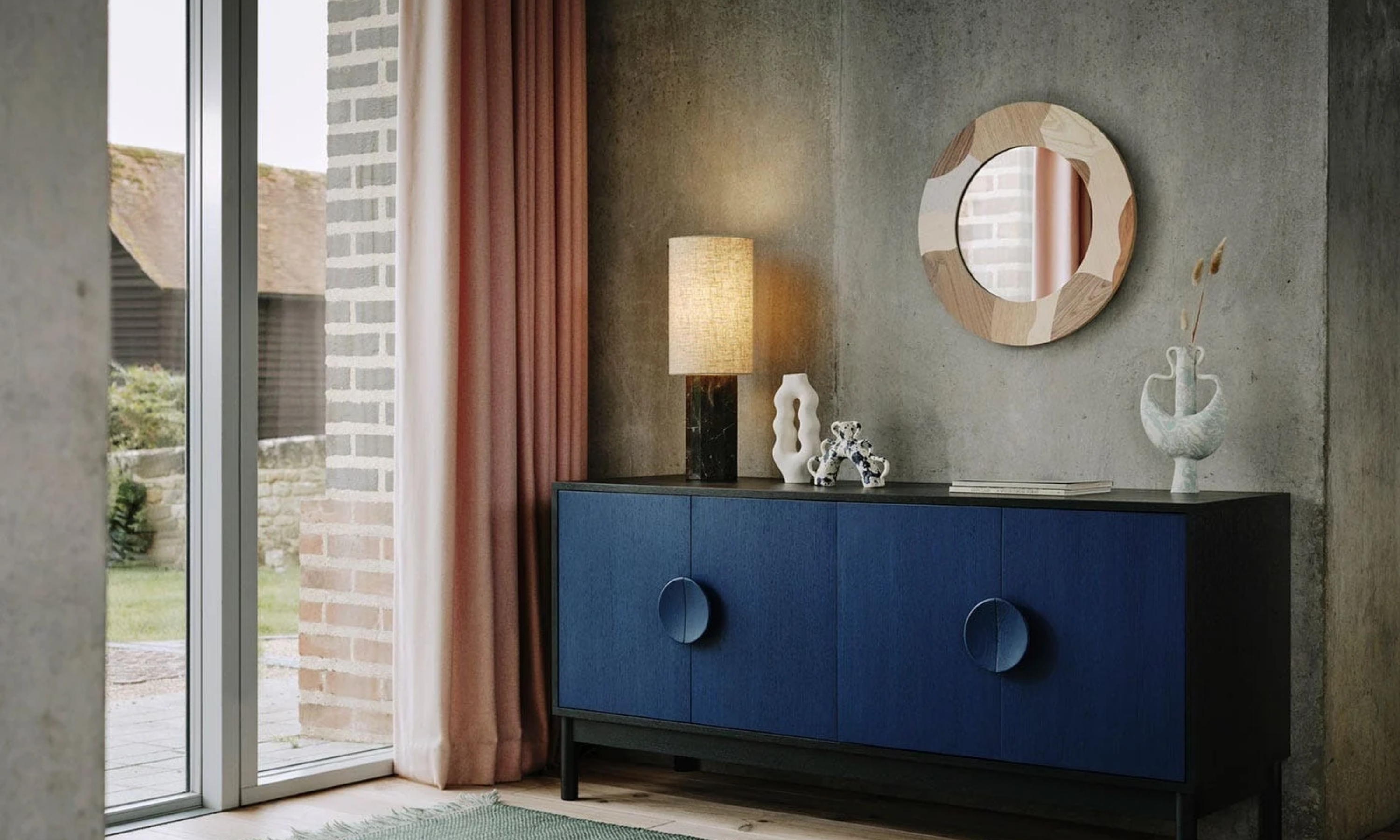 Blue sideboard styled with a linen-shaded table lamp and sculptural ceramics against a textured gray wall, beside soft pink curtains.