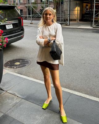 British femal influencer Lucy Williams poses on a NYC sidewalk wearing a white long sleeve flowy top, black woven bag, brown mini skirt, and green Prada square-toe mule kitten heels