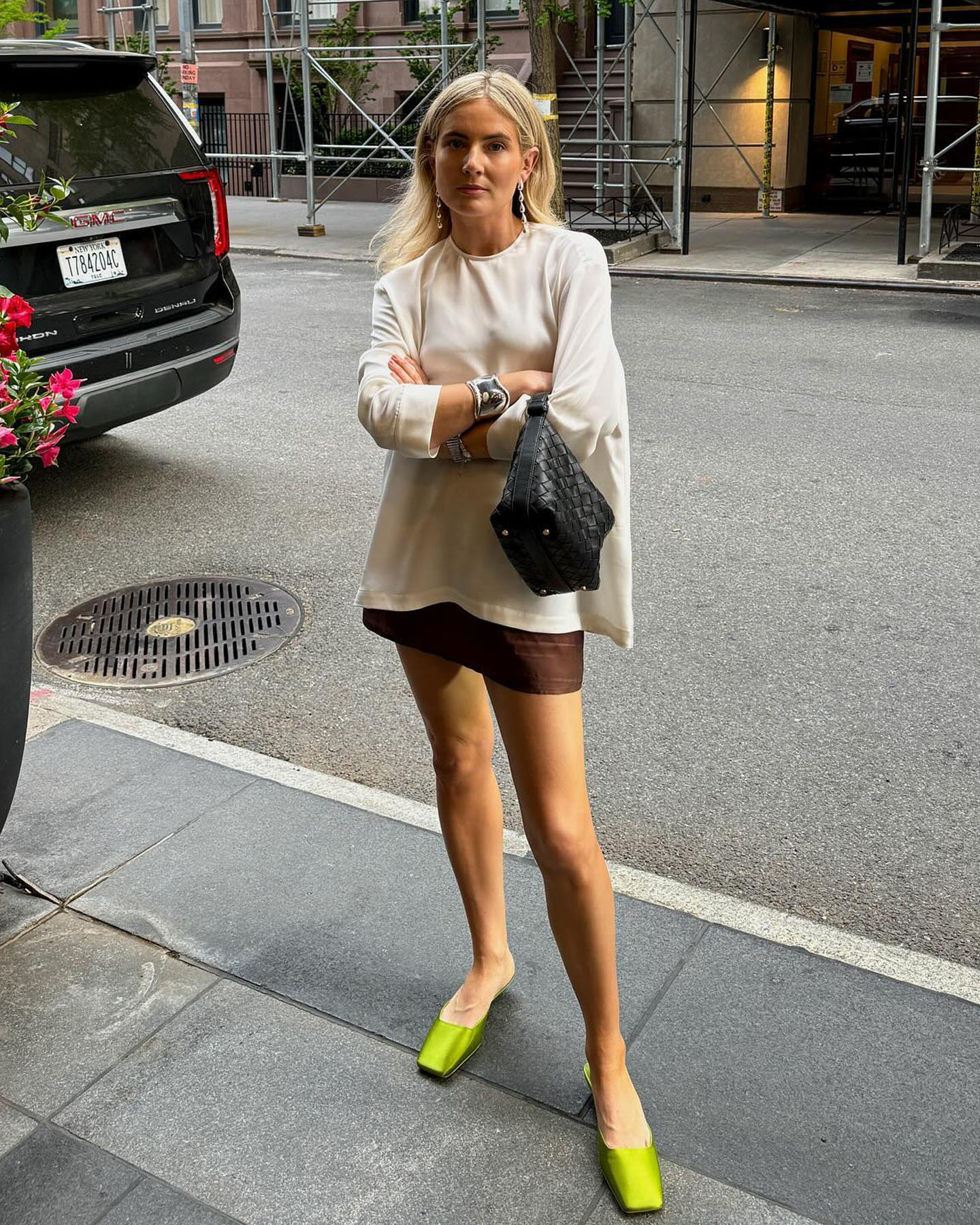 British influencer Lucy Williams poses on a NYC sidewalk wearing a white long sleeve flowy top, black woven bag, brown miniskirt, and green Prada square-toe mules.