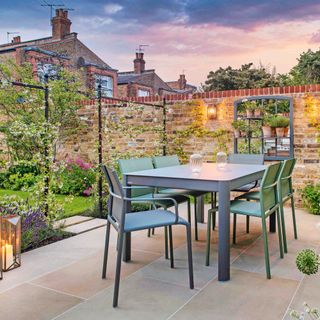 Alfresco dining area in garden with dining set, brass coloured lanterns with tall white wax candles, planter stand, outdoor Critall-style mirrored shelving unit and brick wall decor
